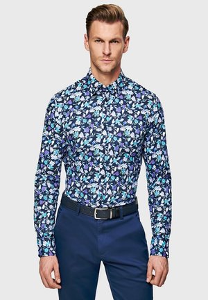 Mężczyzna ubrany w granatową, białą i fioletową koszulę z długim rękawem w kwiatowy wzór, wsuniętą w ciemnoniebieskie spodnie z czarnym paskiem, na jednolitym tle.