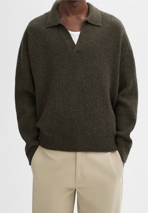 Mand iført en mørkegrøn ribstrikket sweater med krave og beige bukser, hænder delvist i lommerne, neutralt baggrund.