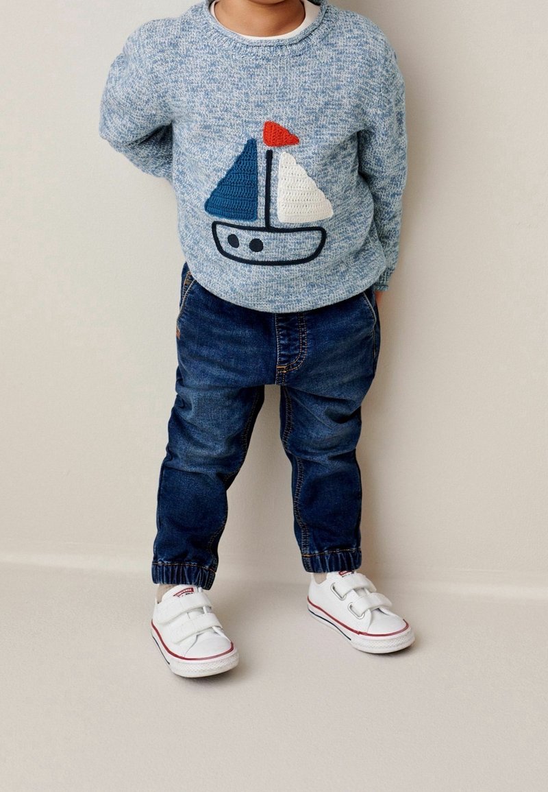 Enfant portant un pull gris avec un voilier bleu et blanc, un jean foncé et des baskets blanches à scratch, debout contre un mur neutre.