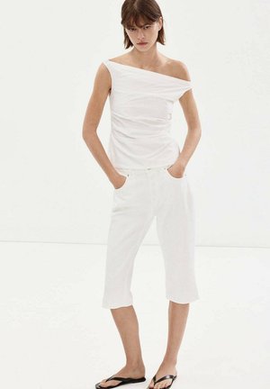 Mujer con top blanco asimétrico sin mangas y pantalones cortos blancos hasta la rodilla con las manos en los bolsillos, de pie sobre un fondo blanco.