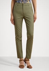 Woman wearing slim-fit olive green pants and brown high-heeled sandals, standing with hands relaxed by sides.