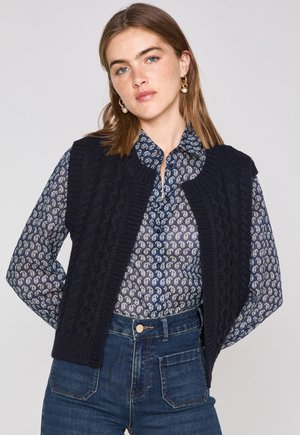 Navy blue knitted vest with cable pattern, round neckline, worn over a blue paisley button-up shirt, paired with dark denim jeans.
