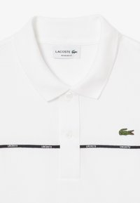 Valkoinen polo-paita teksturoidulla pinnalla, jossa on vihreä brodeerattu krokotiililogo ja mustia raitoja, joissa on "LACOSTE"-teksti. Normaalisti istuva malli.