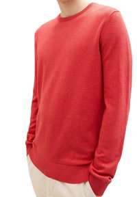 Homme portant un pull rouge uni à manches longues avec col rond, poignets et ourlet côtelés, associé à un pantalon beige clair.