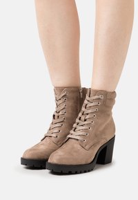 Botines beige de ante con punta redonda, cordones en la parte delantera y cremallera lateral. Presentan un tacón grueso negro y suela texturizada para mayor agarre.