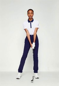 Golf apģērbs, kas sastāv no baltas polo krekla ar tumši zilu apkakli un pogu, kopā ar tumši zilām pieguļošām biksēm un baltām golfu kurpēm, turot golfu nūju.