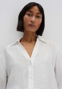 White button-up shirt with a textured fabric, featuring a deep V-neck, long sleeves, and a relaxed fit. Minimalist design accents.