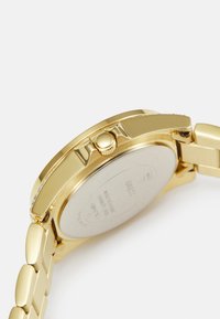 Montre dorée avec un boîtier rond, une couronne texturée et un bracelet à maillons. Présente une finition lisse, un éclat métallique et un design arrière minimaliste.