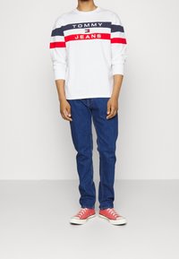 Camicia bianca a maniche lunghe con strisce blu, rosse e bianche, logo "TOMMY JEANS"; jeans blu; sneakers rosse con suola e lacci bianchi.