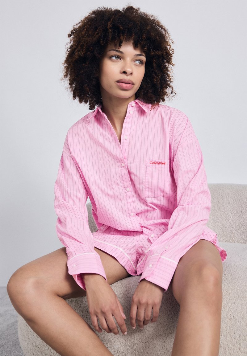 Ροζ, ριγέ, μακρυμάνικη πουκαμίσα με κοντά παντελόνια που ταιριάζουν. Φτιαγμένο από μαλακό ύφασμα, με λευκές λεπτομέρειες και ένα μικρό λογότυπο στο στήθος.