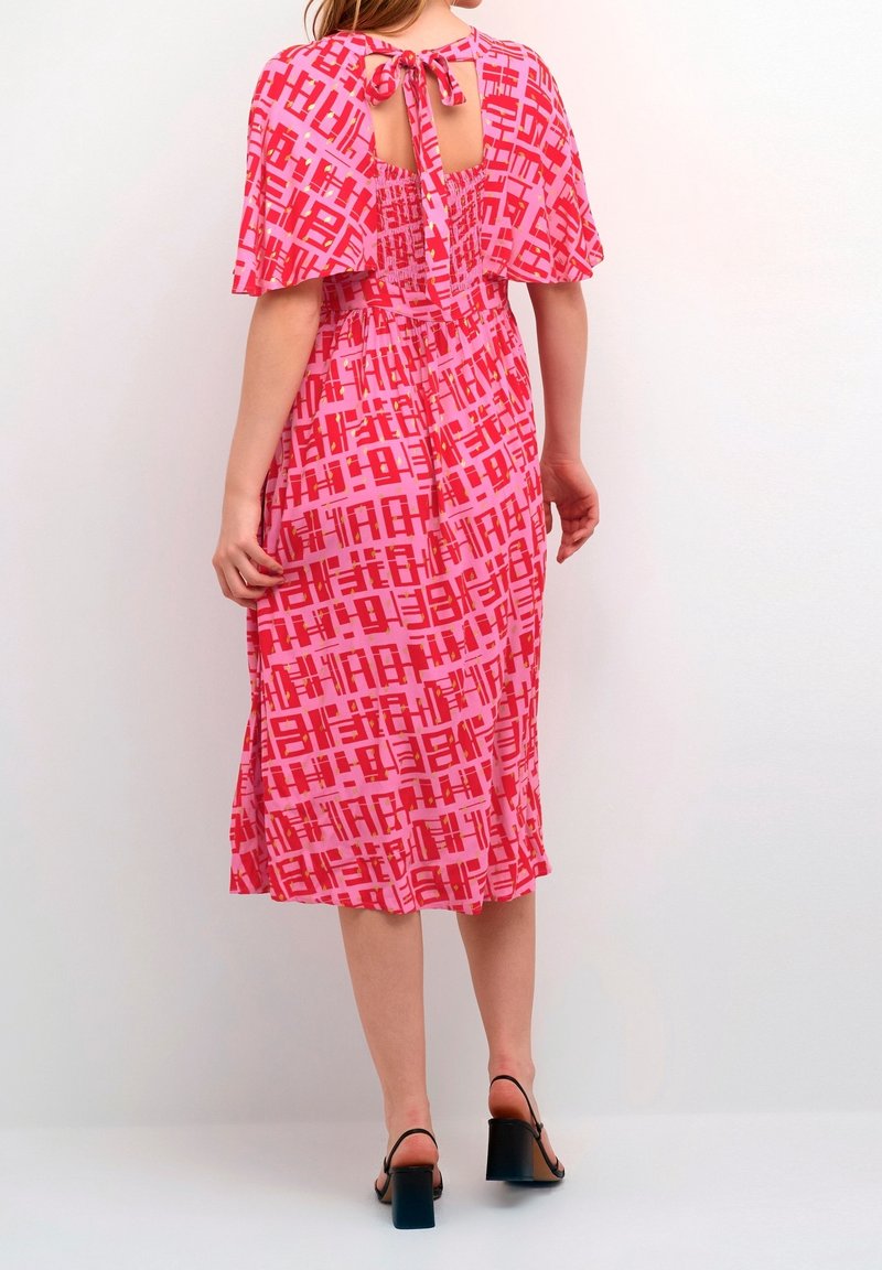 Rotes Kleid mit abstraktem pinken Druck, kurzen flatternden Ärmeln, offener Rückseite mit Bindeschluss und mittellangem Saum. Kombiniert mit schwarzen Blockabsatzsandalen.