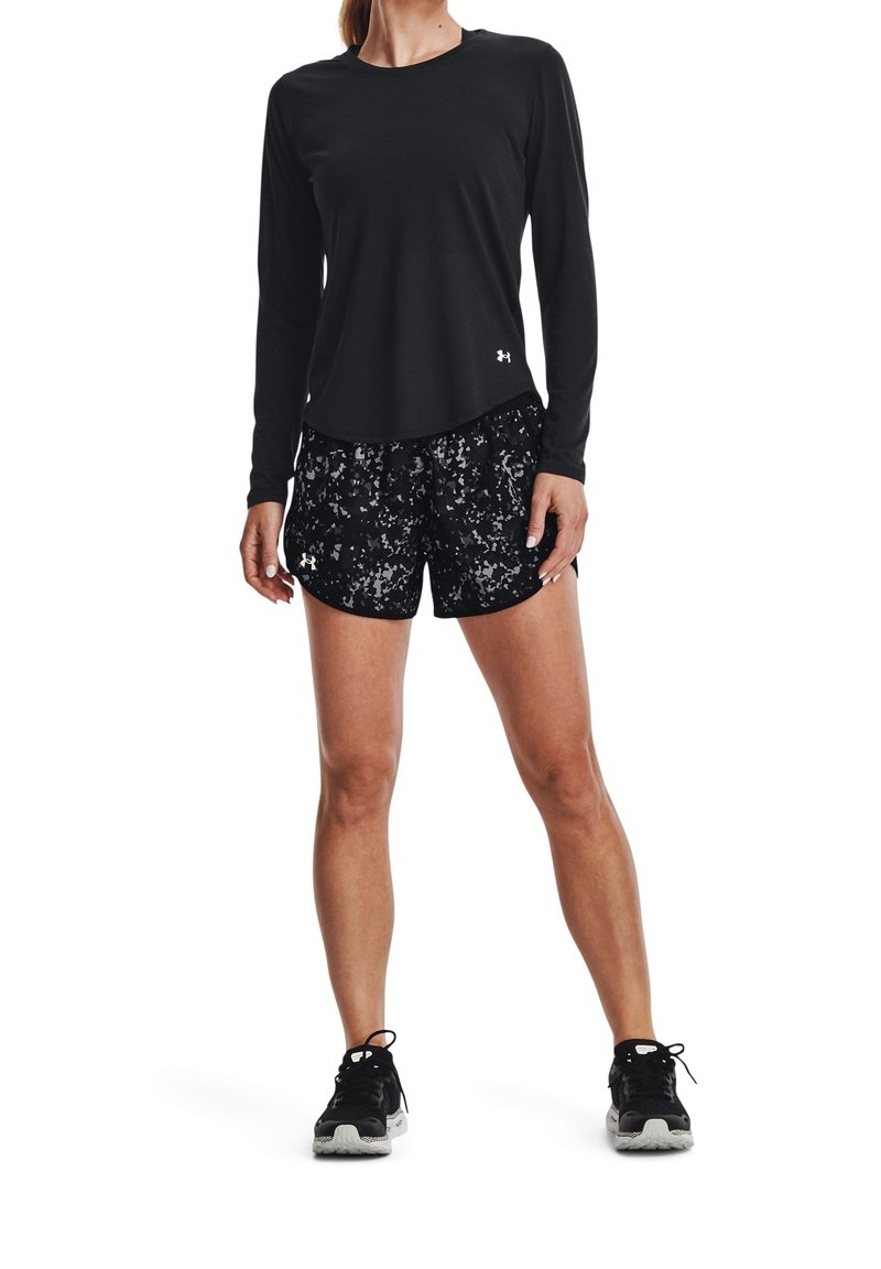 Femme portant un t-shirt noir à manches longues, un short de sport noir à motifs et des chaussures de course noires, debout les jambes légèrement écartées.