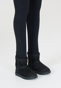 Bottines noires en daim avec un band doux en haut, surface texturée et semelles en caoutchouc, portées avec des leggings noirs.