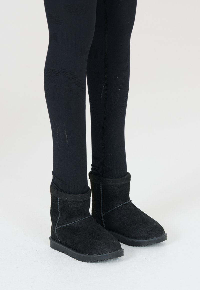 Bottines noires en daim avec un band doux en haut, surface texturée et semelles en caoutchouc, portées avec des leggings noirs.