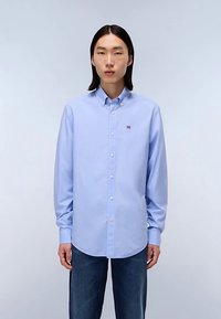 Chemise bleu clair à manches longues avec boutons et col pointu, ornée d'un petit logo de drapeau sur la poitrine et d'une texture de tissu douce et lisse.