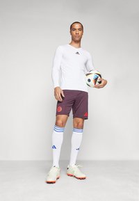 Camiseta blanca de manga larga, pantalones cortos burdeos con logo rojo, calcetas blancas hasta la rodilla con franjas azules y botas de fútbol crema con detalles naranjas.