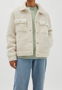 Cremefarvet fleecejakke med to brystlommer, trykknaplukninger og krave. Båret over en lysegrøn sweatshirt og jeansbukser.