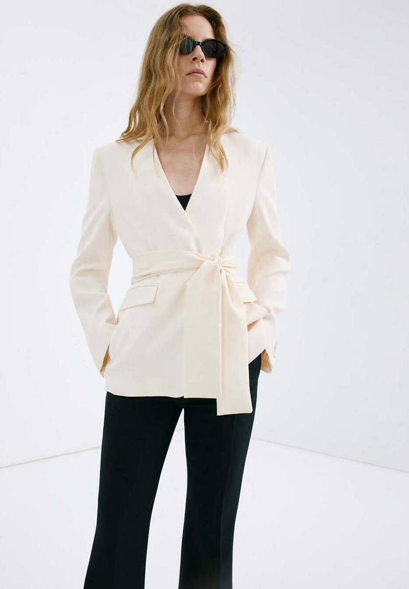Femme portant un blazer beige clair ceinturé, des lunettes de soleil noires et un pantalon noir, debout devant un fond blanc uni.