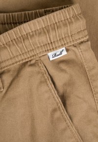 Pantalones color canela con cintura elástica y una pequeña etiqueta blanca de la marca "Reell" cosida cerca de la costura del bolsillo.