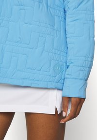 Světle modrá prošívaná bunda má šikmé vzory a texturovaný povrch, doplněná bílou sukní. Na rukávu je viditelná visačka.