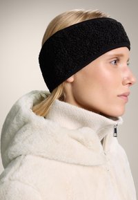 Svart fleece-huvudband med texturerad yta, bärs med en krämfärgad jacka med dragkedja och huva som har en ribbad krage. Slät hudstruktur.