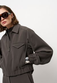 Persoon in een donkergrijze ritsjack met elastische manchetten, schildpadbril en zilveren armband tegen een eenvoudige witte achtergrond.