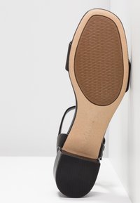 Semelle d'une sandale noire à petit talon, avec une semelle en caoutchouc sur la moitié avant, une semelle intermédiaire lisse de couleur tan et un talon noir strié.