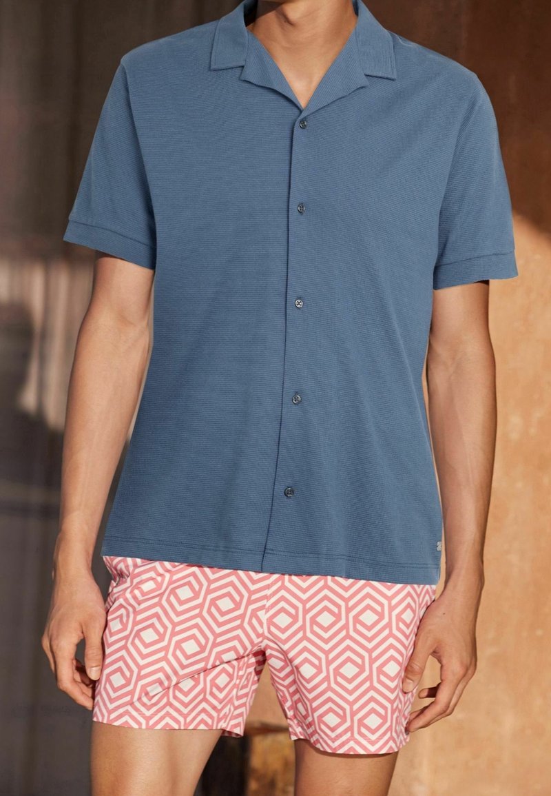 Chemise bleue à manches courtes avec boutons, associée à un short rose à motif géométrique, confectionnée dans un matériau léger avec une coupe décontractée.