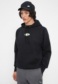Giovane uomo che indossa un cappellino Nike nero e una felpa nera con un piccolo logo chiaro, abbinati a pantaloni grigi con una toppa Nike.