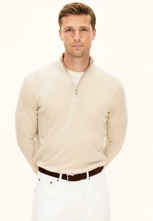 Uomo con capelli castano chiaro che indossa un maglione beige con cerniera fino a metà collo, pantaloni bianchi e cintura marrone scuro, in piedi con le mani dietro la schiena.