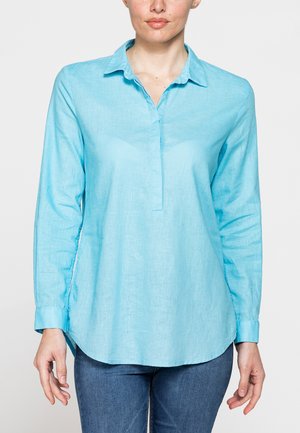 Donna che indossa una camicetta azzurro chiaro a maniche lunghe con colletto abbinata a jeans blu, mostrata dal collo ai fianchi su uno sfondo semplice.
