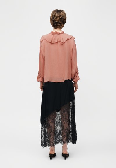 Personne portant un chemisier rose transparent à col froufrouté et une jupe asymétrique noire avec un voile en dentelle, vue de dos.