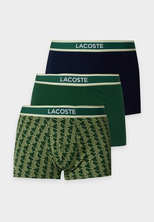 Skjortlös man som står barfota iförd mörkgröna Lacoste boxer shorts mot en enfärgad vit bakgrund.