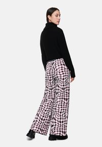 Frau in einem schwarzen Rollkragenpullover, rosa und schwarz karierten weiten Hosen und schwarzen klobigen Stiefeln, die steht und über ihre Schulter schaut.