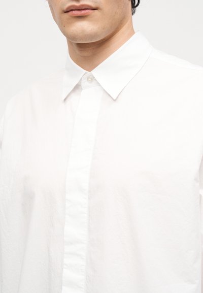 Άνδρας με λευκό πουκάμισο με κουμπιά και μυτερό γιακά, που δείχνει τον λαιμό και το κάτω μέρος του προσώπου σε απλό φόντο.