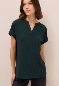 Mørkegrøn kortærmet bluse med V-hals og tekstureret stof, der har diskrete lodrette linjer. Løs pasform med et afslappet design.