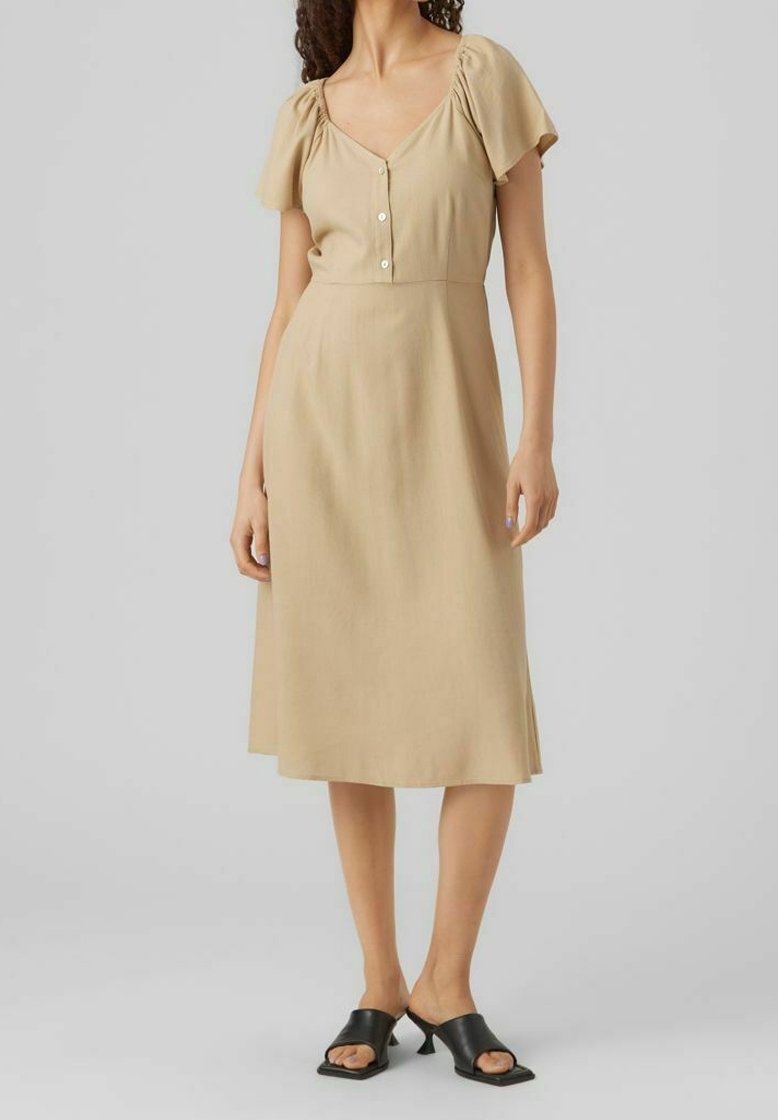 Femme portant une robe midi beige à manches courtes bouffantes, col en V avec boutons, associée à des sandales à talons noires et ouvertes.