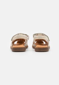 Sandalias abiertas de color beige claro con correas ajustables, plantilla acolchada y suelas de goma marrón, con detalles recortados en los laterales.