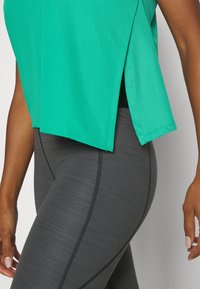 Grön nät-träningslinne med sidoklyftor, ihop med strukturpäron mörkgrå leggings med framträdande sömmar och en figurnära design.