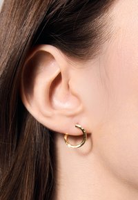 Złota kolczyk w kształcie koła o młotkowanej fakturze, z otwartym designem. Noszony na uchu, podkreślający płatek ucha i sąsiadujące włosy.