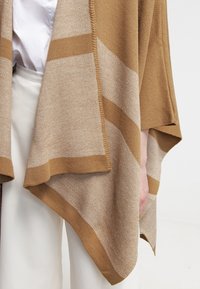 Bruin en beige gebreide poncho met geometrische patronen, open voorkant en wijde mouwen, met een gestructureerd weefsel en contrasterende accenten.
