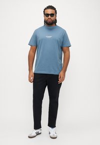 Blå bomuld t-shirt med korte ærmer og et logo på brystet, kombineret med sorte bukser og hvide sneakers med sorte striber.