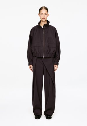 Czarna krótka kurtka z wysokim kołnierzem i zapięciem na zamek, w połączeniu z szerokimi spodniami o asymetrycznym układzie. Gładka faktura tkaniny.