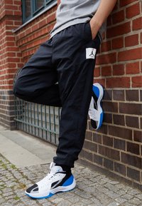 Person som har svarta byxor med en vit Jumpman-logotyp lutar sig mot en tegelvägg, iförd högskor i vitt, svart och blått.