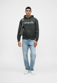 Felpa nera in cotone con tasca frontale e cordino, con logo "Calvin Klein". Abbinata a jeans in denim blu chiaro e sneakers bianche.