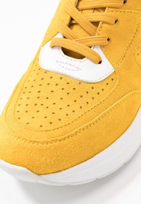Yellow suede sneaker toe with perforations, white leather accent, yellow laces, and white rubber sole on a white background.