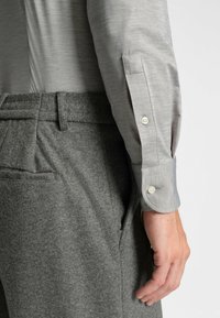 Camicia grigia a maniche lunghe con una sottile texture, polsini con bottoni, abbinata a pantaloni grigi in misto lana con etichetta del logo e tasche laterali.