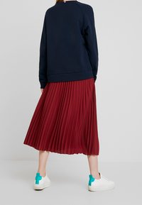 Marinblå sweatshirt med raglanärm, matchad med en röd plisserad midikjol. Vita sneakers har turkosa detaljer på hälen.