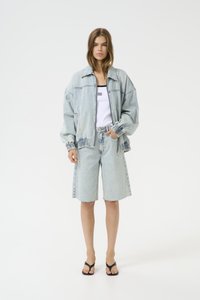Šviesiai mėlynas džinsinis oversize švarkas su elastiniais rankogaliais, derinamas su aukšto talių šortais. Modelis dėvi baltą marškinėlį be rankovių ir juodas šlepetes.