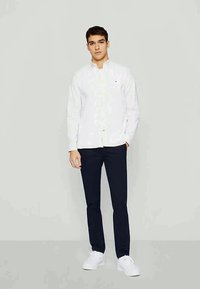 Camisa branca de manga longa, com botões e um pequeno logótipo, combinada com calças azul-marinho e ténis brancos. Textura suave e ajuste cuidado.